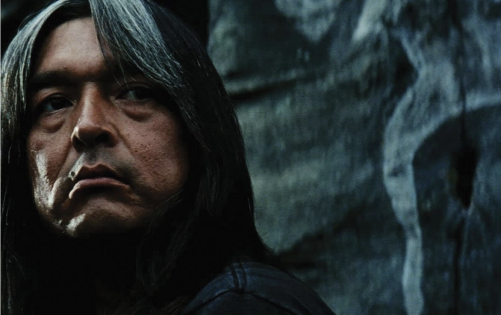 A close-up shot of actor Graham Greene, who has black hair with greying bangs. He is standing in front of a rock wall.